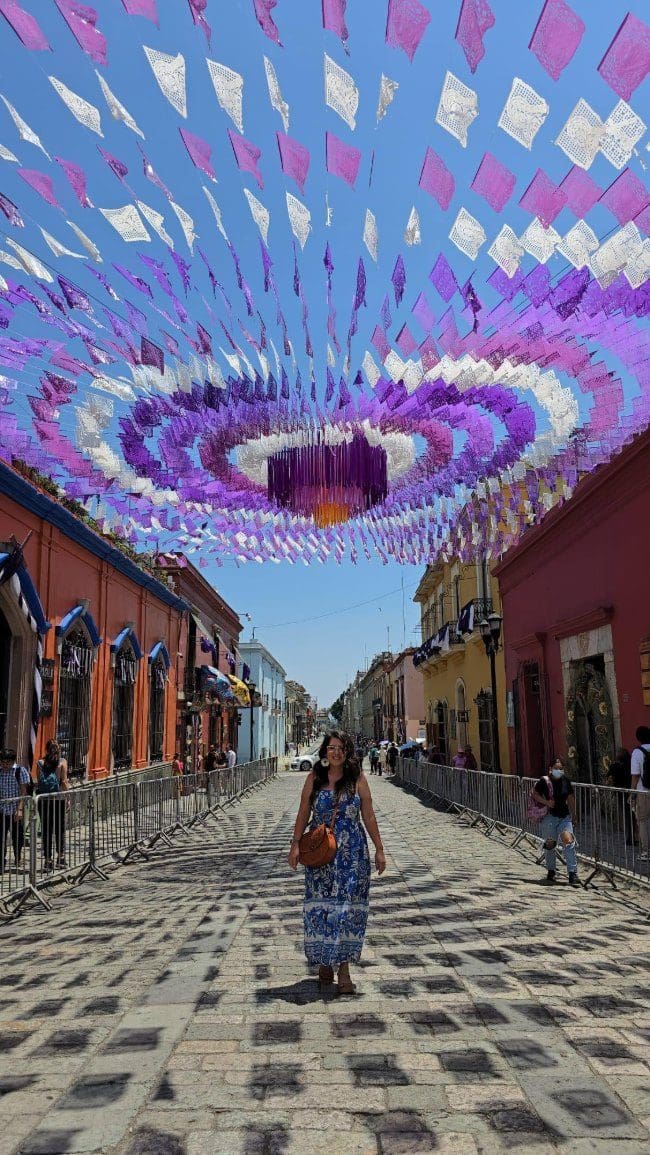 Woman in a blue floral dress standing under a purple papel picado canopy on a colorful Oaxaca City street — Oaxaca honeymoon inspiration
